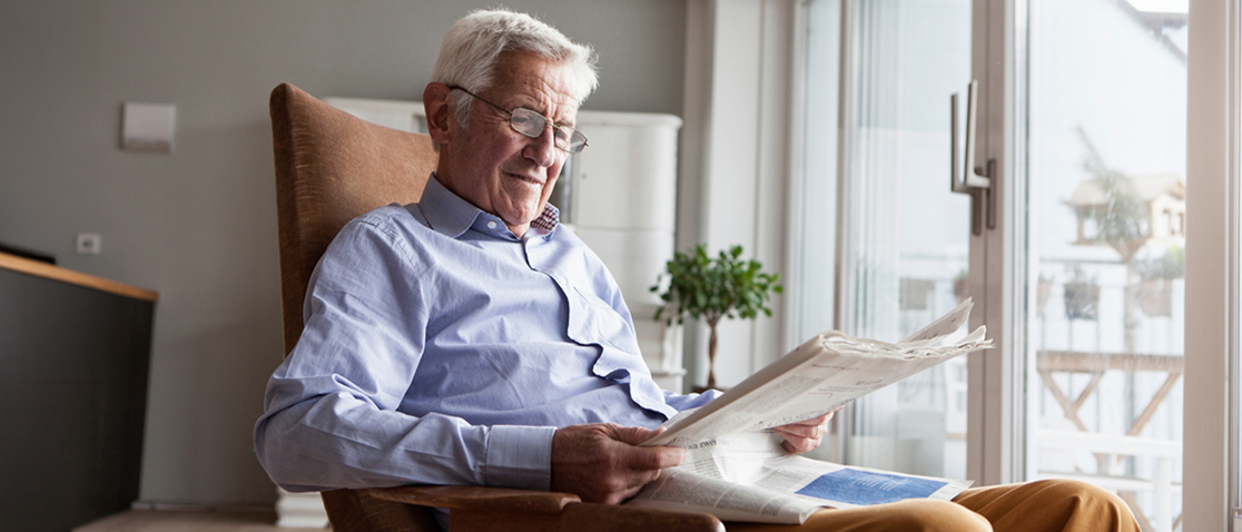 A white-haired man wearing glasses, a blue button-up shirt, and dark khaki pants smiles while reading the newspaper in a rocking chair in front of his home’s wall-length windows and glass patio doors.