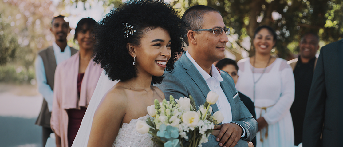 A young woman wearing a white sleeveless wedding gown, dangling silver earrings, and an intricate silver hairpin smiles while holding onto the arm of a man wearing a muted teal suit that matches the white and teal bouquet in the bride’s hands, with a smiling crowd looking on.