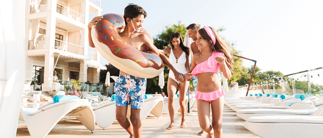 A laughing man, woman, boy, and girl all wearing swim suits run excitedly through rows of white outdoor lounge chairs at a large resort on a sunny day.